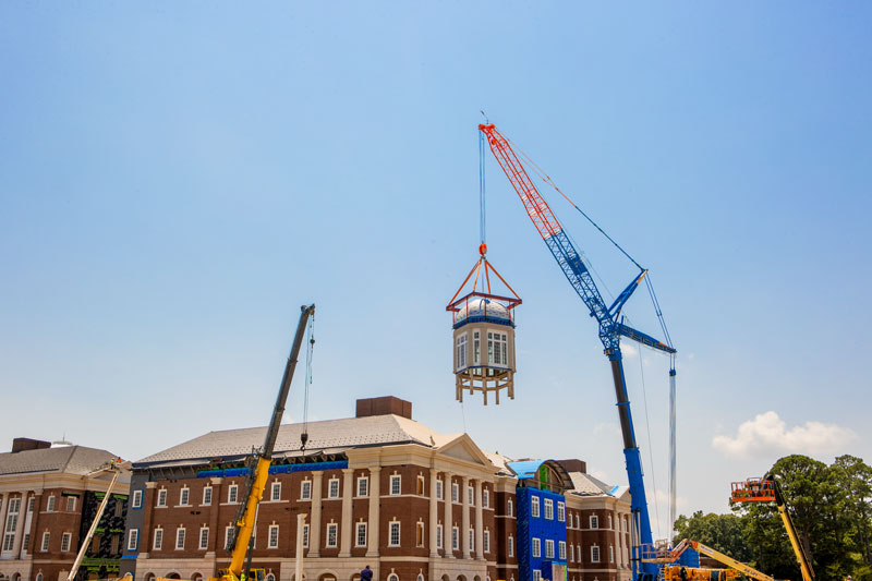 A crane lifts a cupola to the top of a building that is under construction