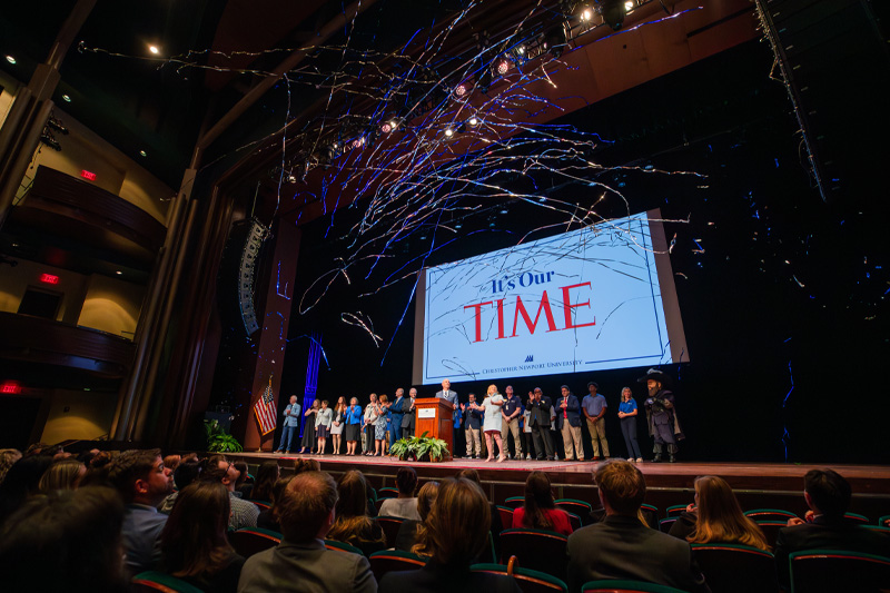 A crowd of university student-athletes, faculty and staff stand on the stage with Christopher Newport President William Kelly and his wife Angie
