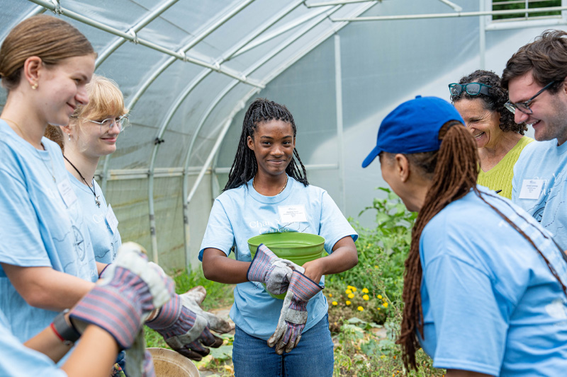 Students wearing light blue Day One of Service shirts put on gardening gloves while standing in a greenhouse