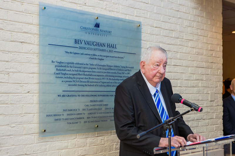 Bev Vaughan stands in front of a plaque bearing his name on a wall inside of the newly named Bev Vaughan Hall.