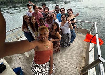 Group selfie of about fifteen smiling young people on the deck of a boat, with a wide river and green trees in the background.