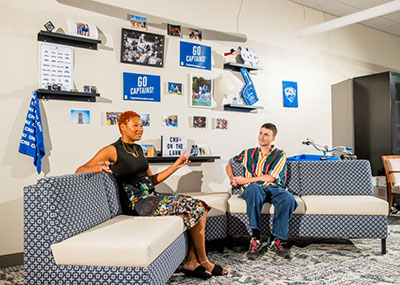 Two people sit on a blue and white patterned sectional couch in a room decorated with CNU memorabilia, including posters that say GO CAPTAINS! and a sign that says CNU ON THE LAWN. The woman on the left is speaking to the man on the right.