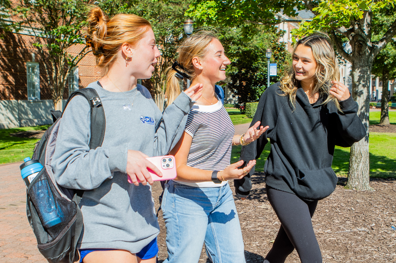 Three smiling female college students walk and talk outside on a sunny day. The student on the left wears a grey sweatshirt and has a backpack. The s