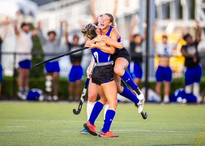 Two female field hockey players in blue and black uniforms embrace and cheer after a goal, with one jumping into the arms of the other. Spectators with their arms raised are visible in the blurred background.