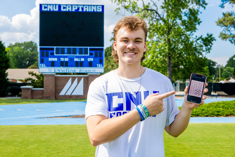 A young man wearing a white shirt with CNU FOOTBALL is standing in front of the scoreboard at TowneBank Stadium holding a smartphone.