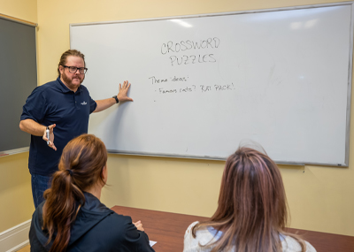 A bearded man wearing glasses and a blue polo shirt is teaching a class, standing next to a whiteboard that has CROSSWORD PUZZLES written on it, along with the theme ideas Famous cats? and RAT PACK!. Two students with their backs to the camera are seated at a table.