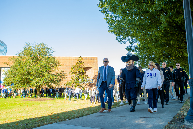 CNU mascot, a man in a suit, and a woman in a CNU sweatshirt walking along a paved path outdoors.