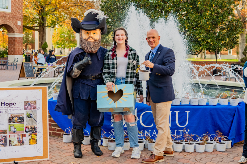 A mascot dressed as Captain Christopher Newport, a young woman, and an older man in a suit—stand in front of a fountain and a blue table.
