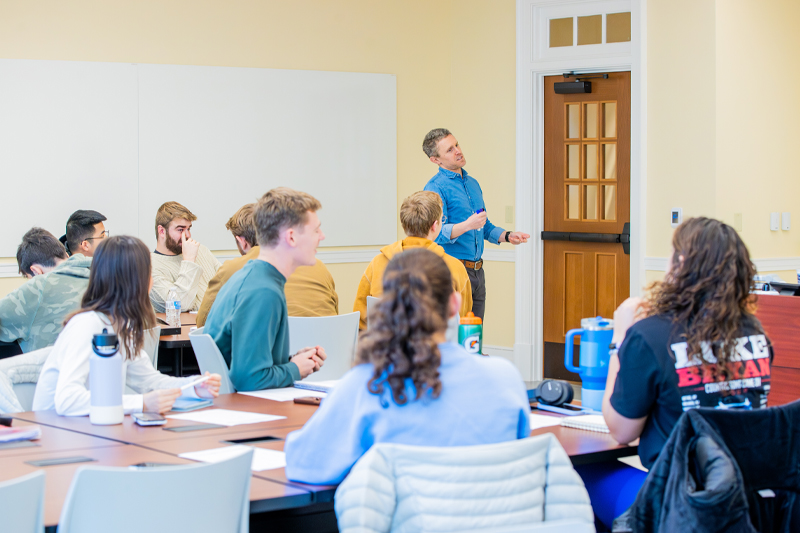 A wide shot of a college classroom where Professor Matthew Morena is speaking and gesturing to a diverse group of students seated.