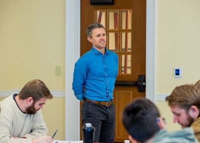 Dr. Matthew Morena wears a blue button-down shirt and dark pants stands smiling in front of a closed wooden door with glass panels in a classroom. Students are seated in the foreground, writing or looking down.
