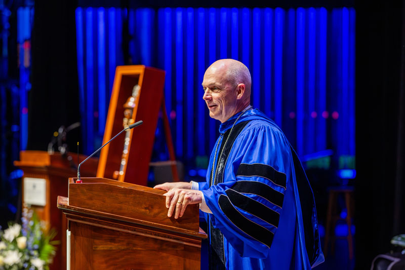 CNU President William G. Kelly wears a bright blue academic robe with black trim stands behind a wooden podium, speaking into a microphone. 
