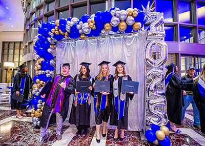 Four graduating students in black caps and gowns pose for a photo under an archway decorated with blue, silver, and gold balloons. Three of the students are holding their diplomas. To the right of the group, a large silver balloon arrangement spells out 2025.