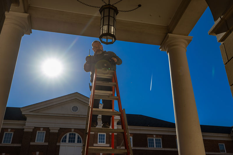A low-angle silhouette of a maintenance worker on a ladder, working on an outdoor hanging lantern under a bright, sunny sky. 