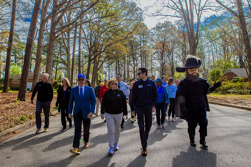 President William Kelly leads a group of people, including campus officials and Captain Chris, walking down a tree-lined residential street.