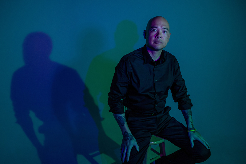 Dr. Sherman Lee sits on a stool against a teal wall. Dramatic blue and purple lighting casts a large shadow behind him.
