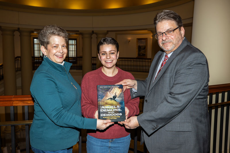 Three authors smile while holding their book, Angels, Demons, and Demigods, in a building with columns and a balcony.