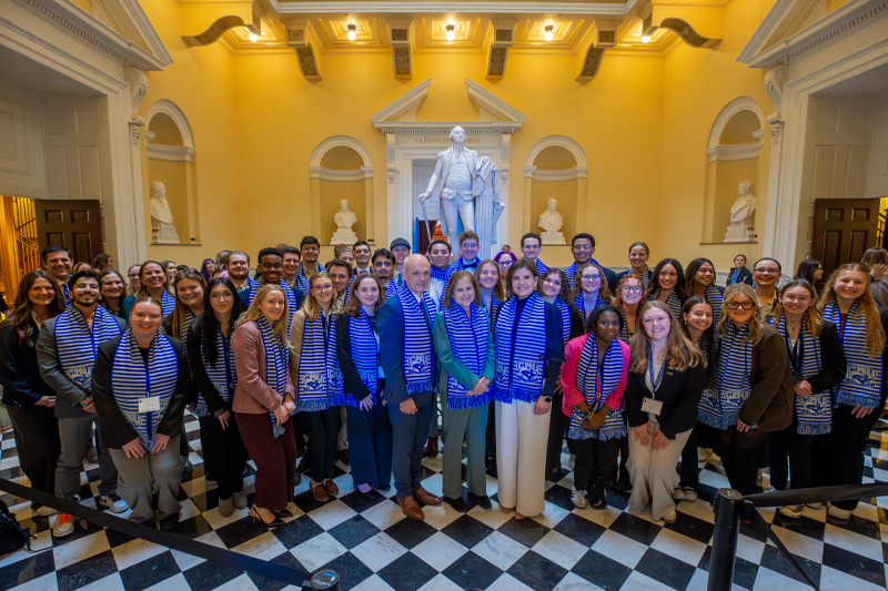 A large group of students in blue striped scarves poses with leaders in a grand, domed hall with a statue.