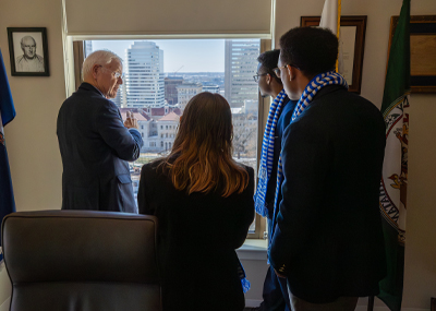 An older man gestures toward a city skyline while talking to three students by an office window.