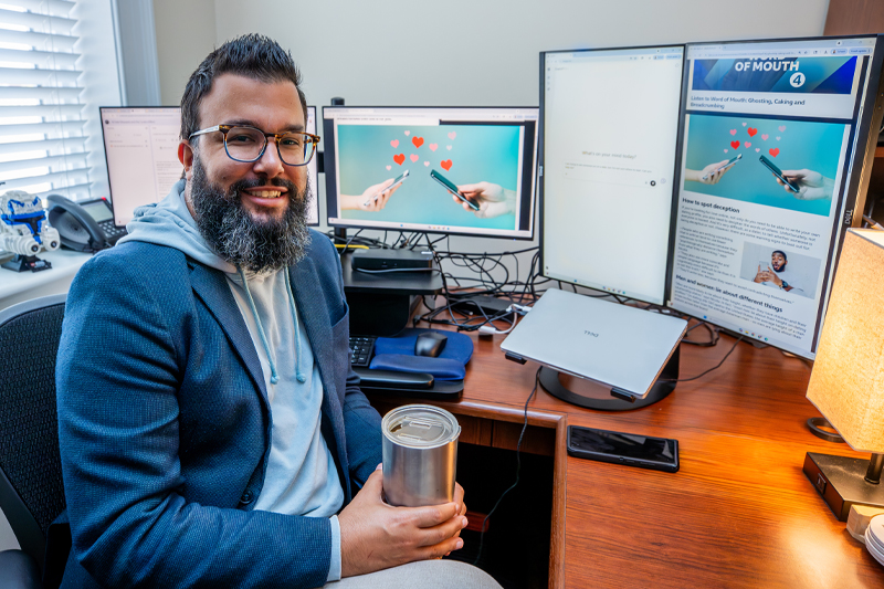 Dr. David Roache, in a blazer sits at a desk with four monitors displaying articles about AI, dating, and deception.