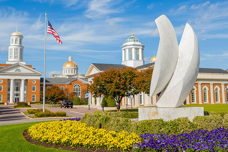 The entrance of campus with a statue in the foreground and a flagpole with the American flag, the Trible Library and Pope Chapel in the background.