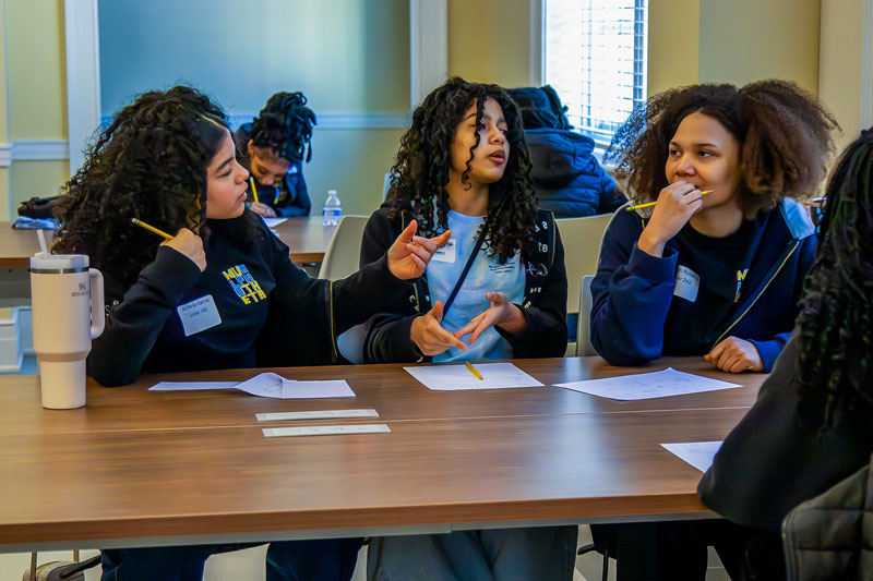 Three students with dark curly hair sit at a long table, discussing work with pencils and paper at a competition.