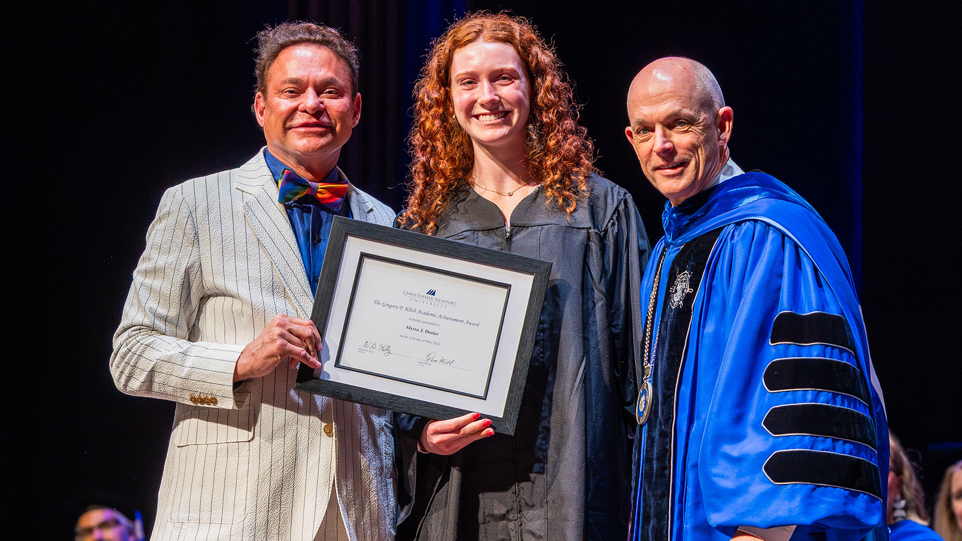A graduate with long curly hair holds a framed certificate between Greg Klich and CNU President Bill Kelly on stage.
