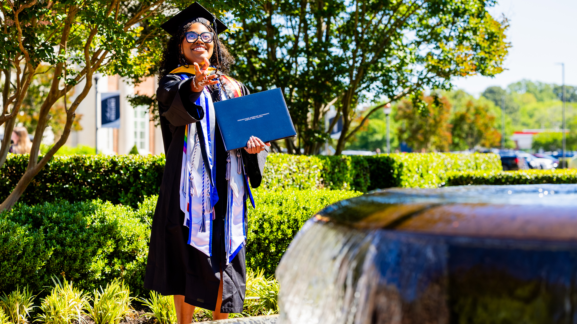 A student in graduation regalia holds onto their diploma and tosses their penny in the fountain.