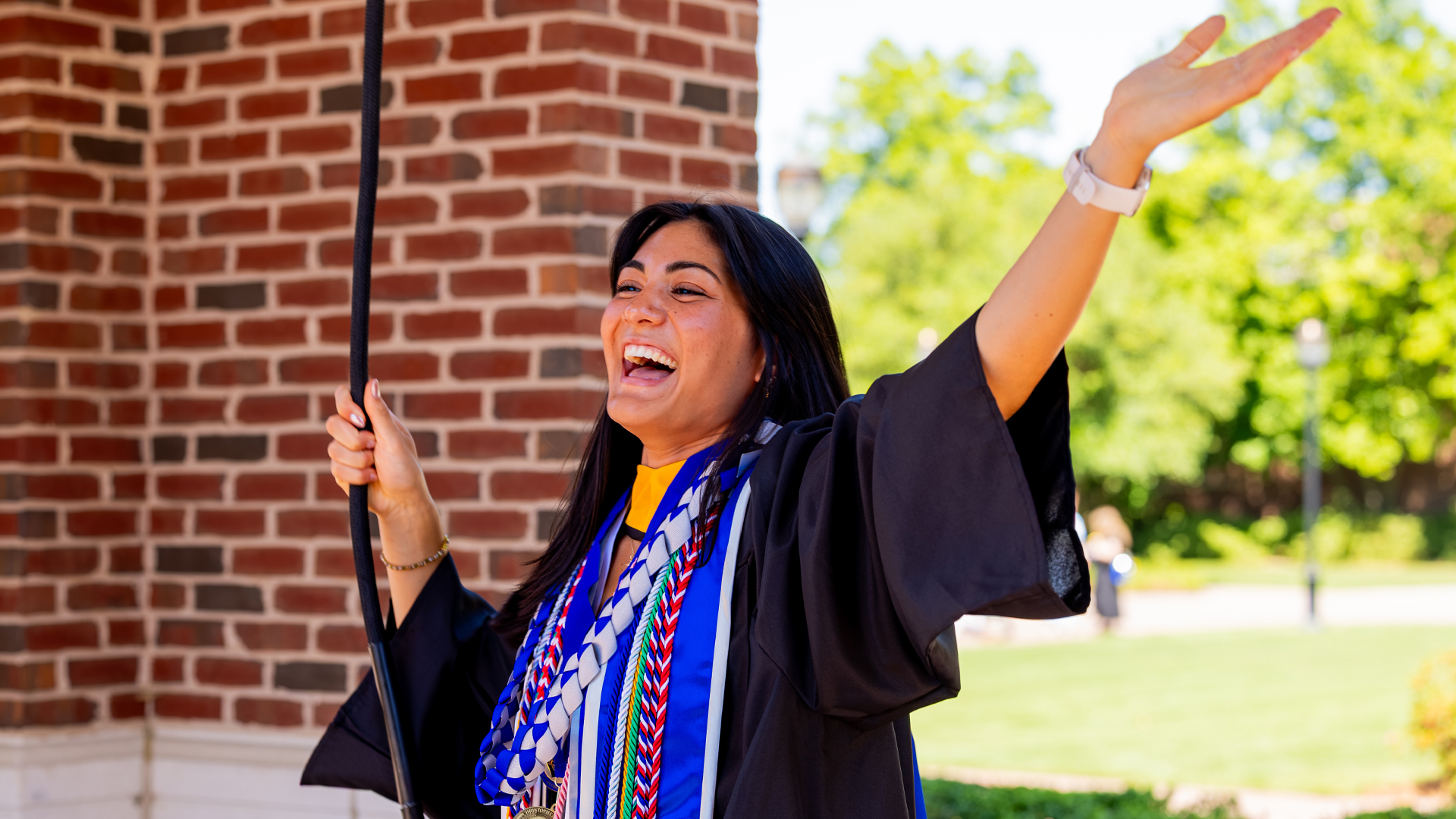 A student in graduation regalia rings the bell and extends her arm and has a smile on her face.
