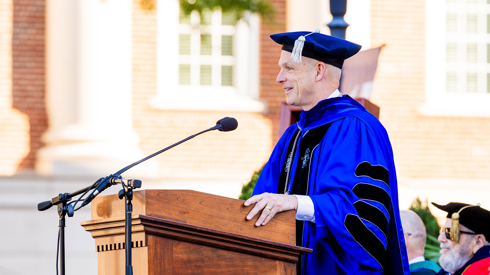 CNU President Bill Kelly, in a blue doctoral robe stands at a podium