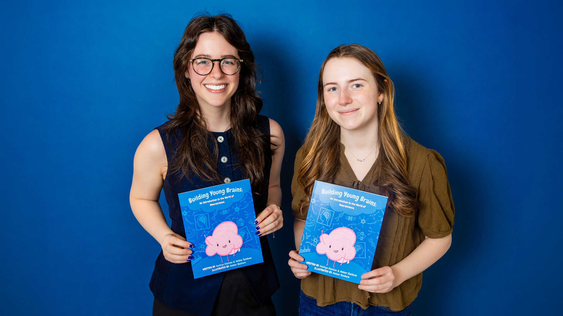 Amber Baldwin and Kaitlyn Kinslow hold copies of their childrens book against a blue backdrop.