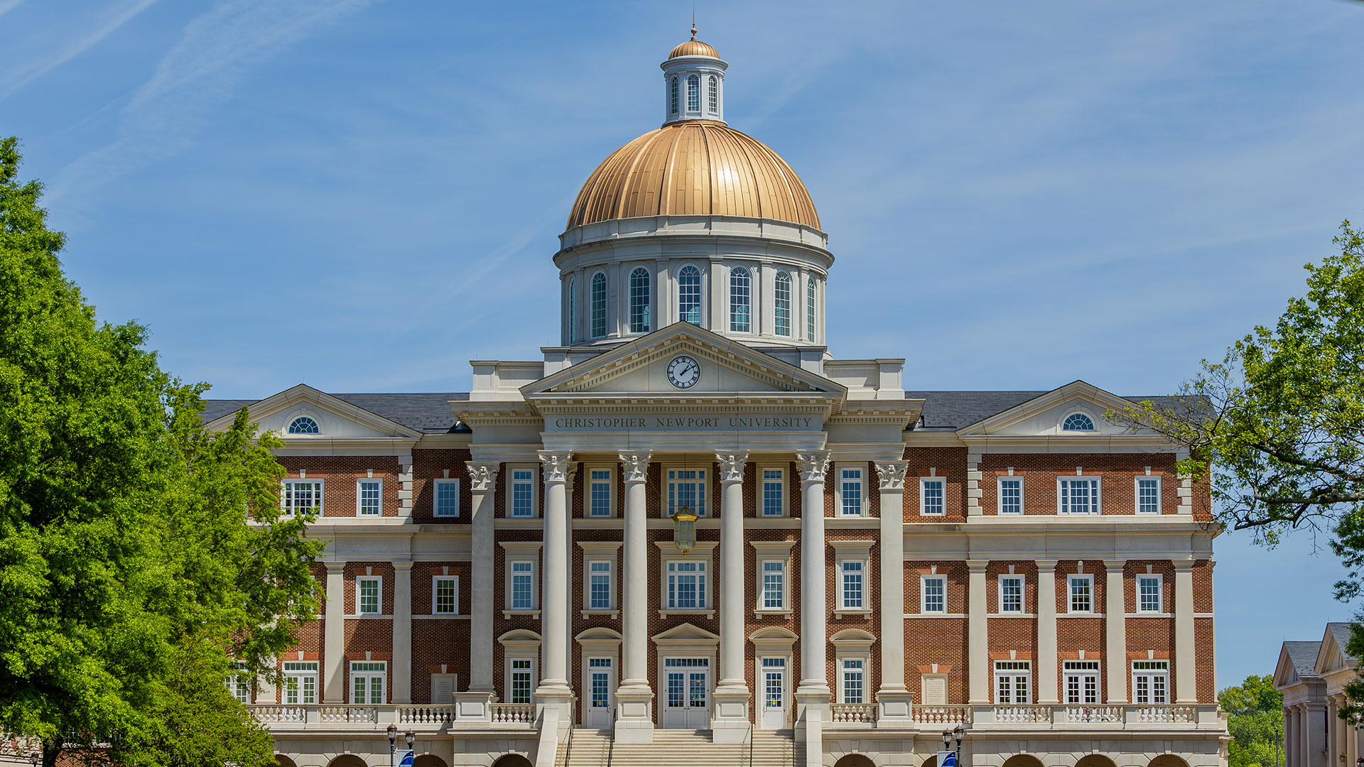 Christopher Newport Hall, a grand, classic university building with a gold dome and white columns, surrounded by a green lawn and trees with autumn foliage under a bright blue sky.