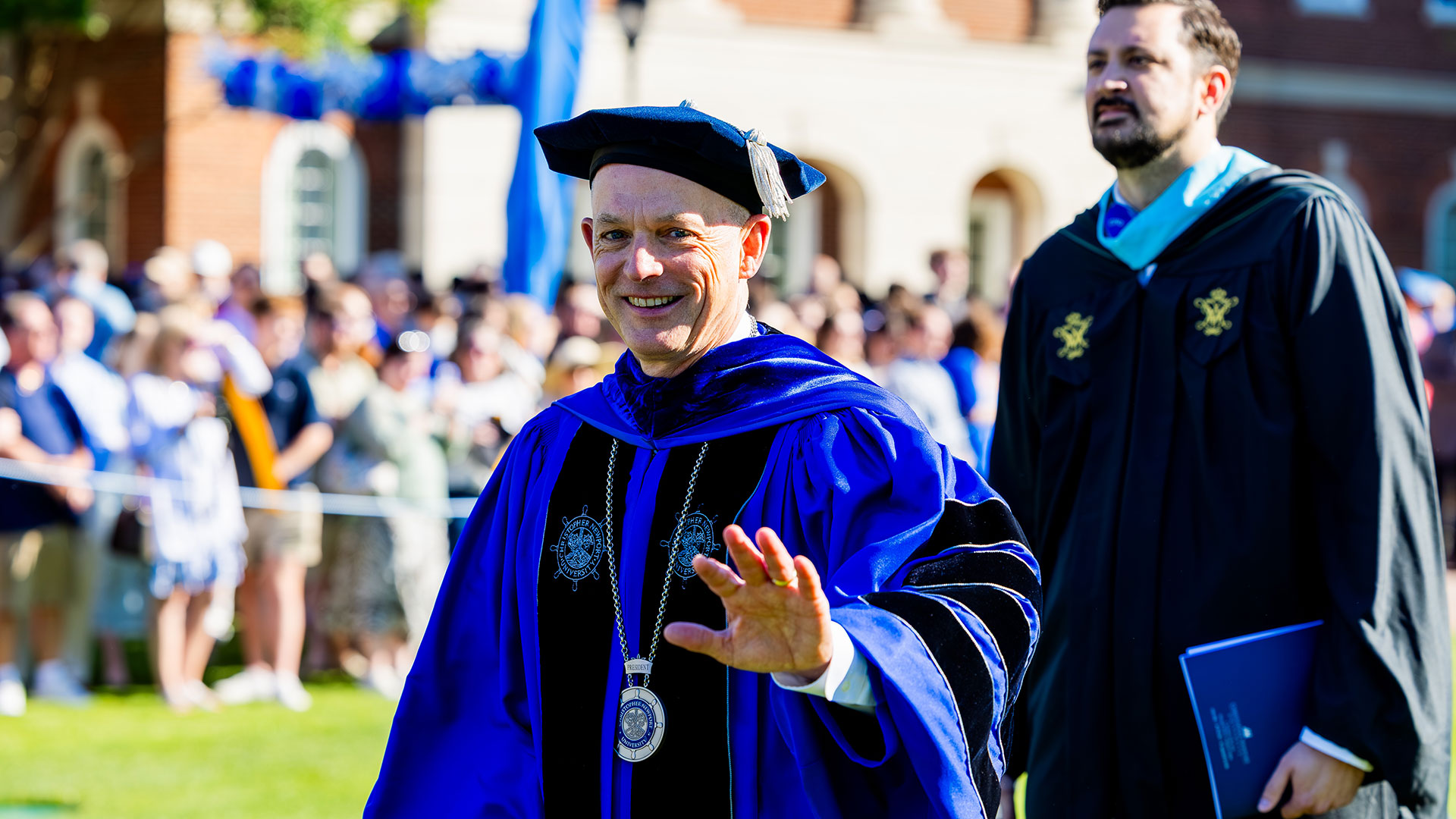 CNU President Bill Kelly waves at the camera while wearing commencement regalia.
