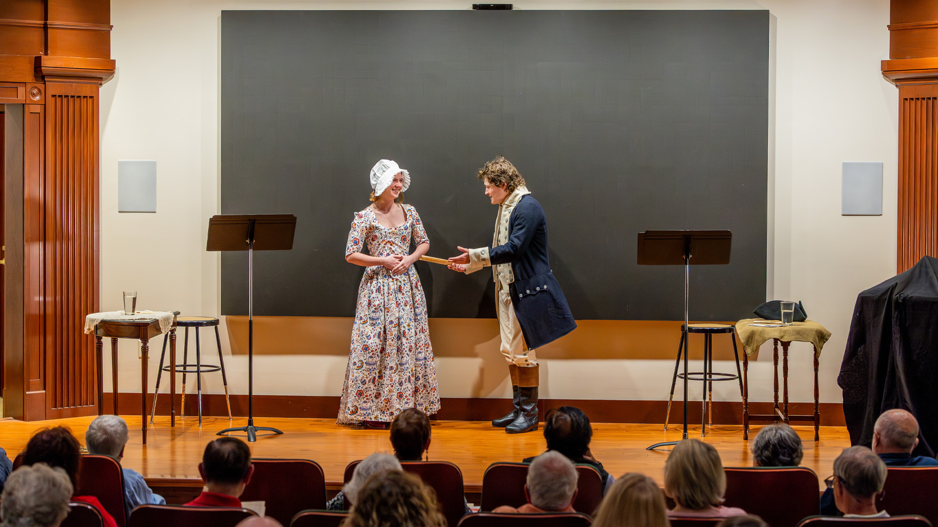 A female student, left and a male student, right wear Revolutionary era garb while standing on a stage.