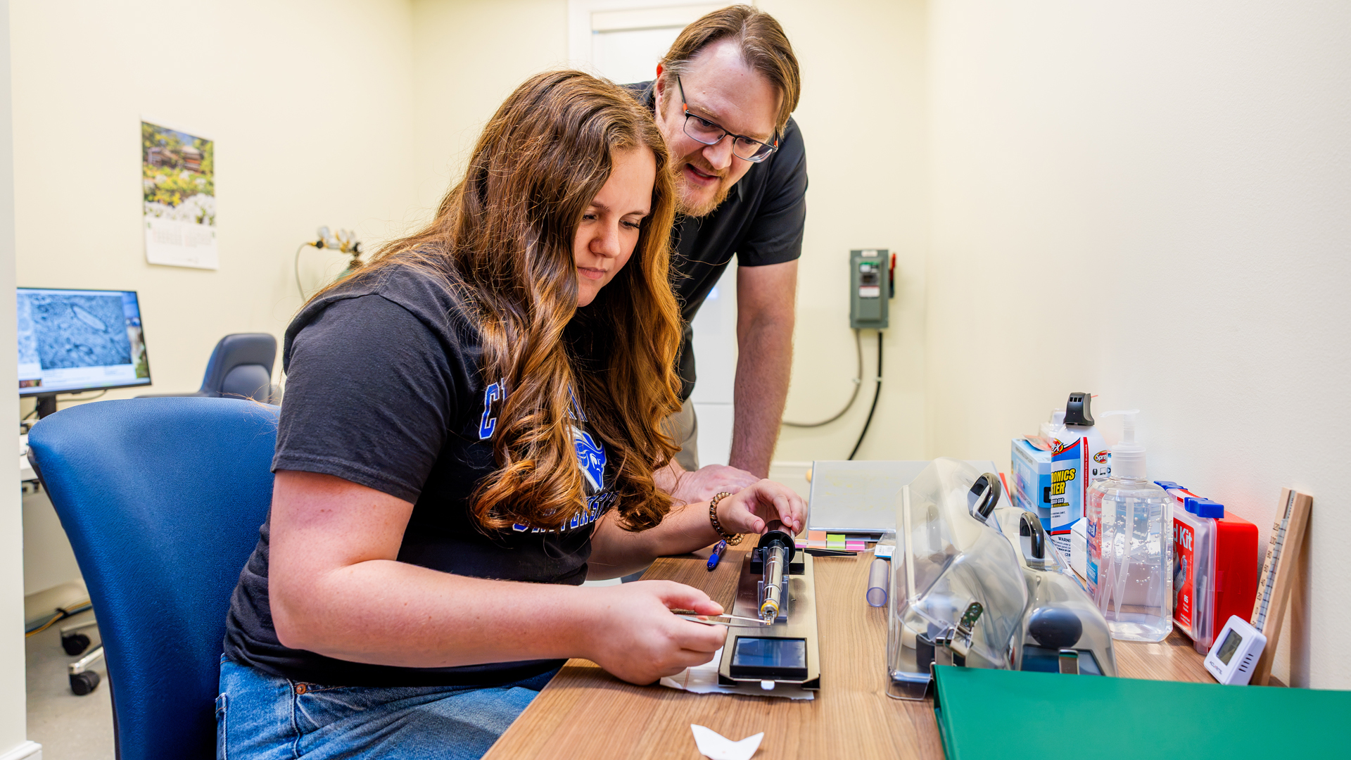A student holds tweezers in front of a piece of lab equipment while a professor looks on.