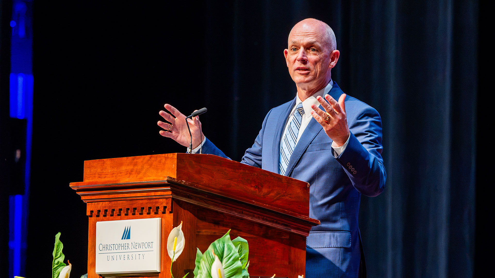 CNU President Bill Kelly stands at a podium with hands wide as he addresses an audience.