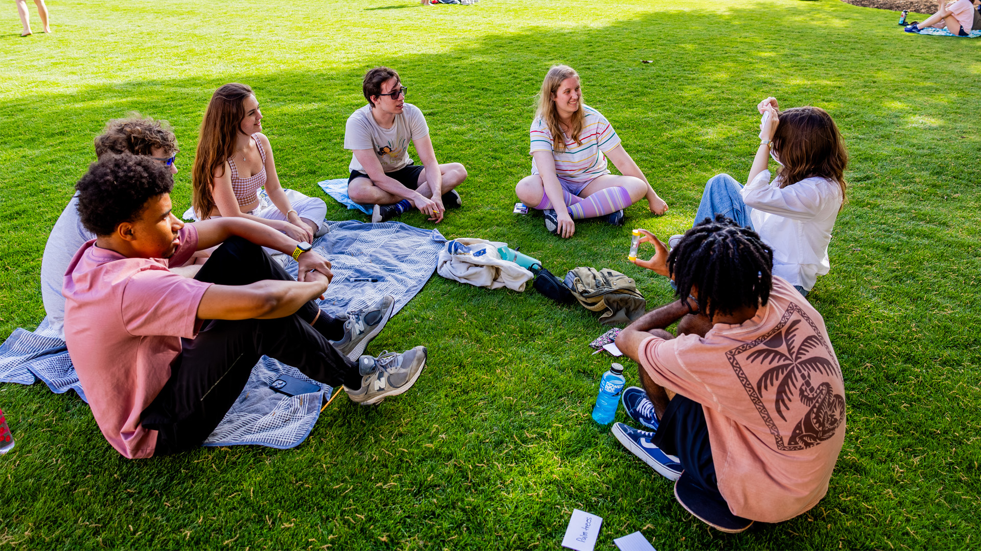 A group of students sit in a circle outside on a green lawn.