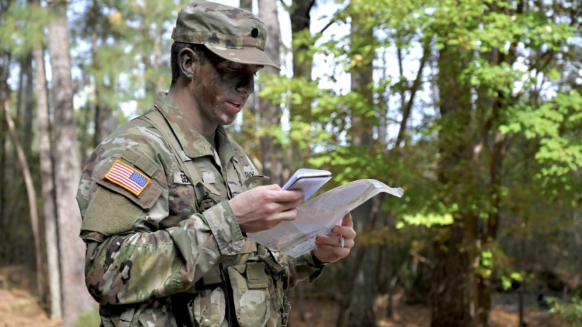 A student in army fatigues and face paint looks at a map and a notebook while in the wilderness