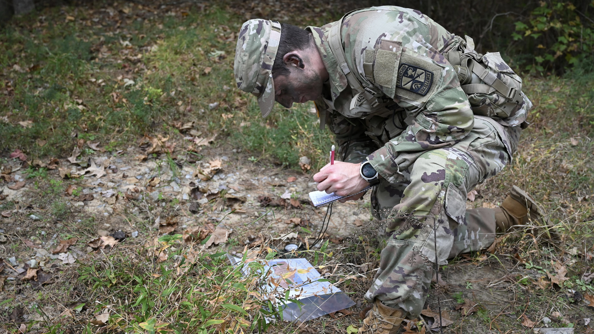 A student wearing army fatigues kneels on the forest floor while looking at a map and writing on small notepad