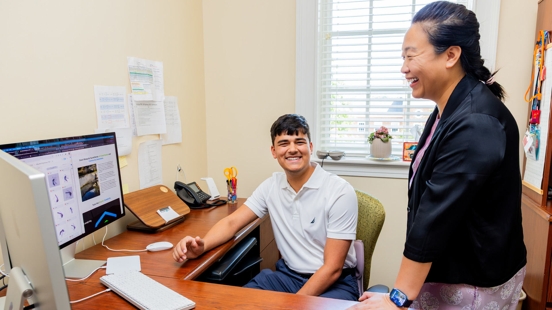 A student sits at a desk in front of a computer screen smiling with a professor standing beside him.