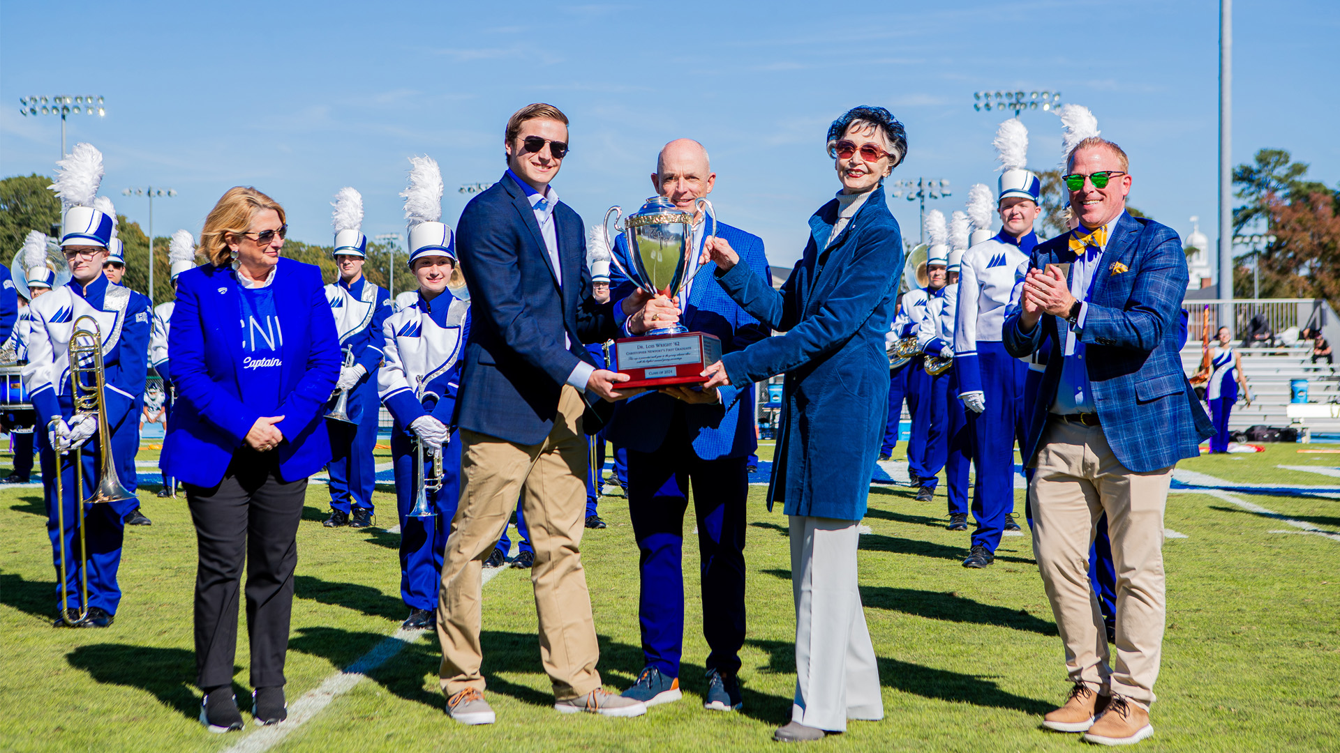 Three individuals hold a silver trophy on a football field with a marching band behind them.