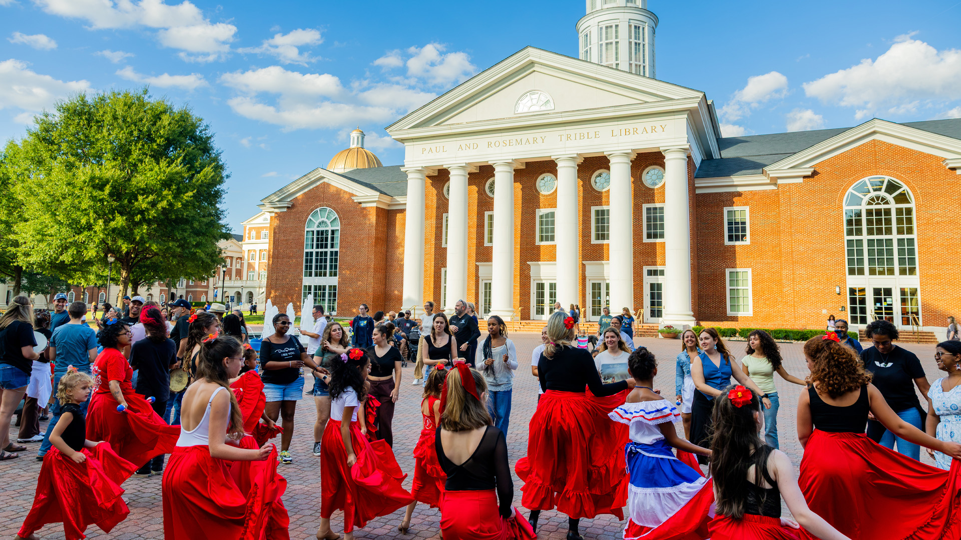 A group of individuals stand in the plaza, some in red skirts to dance.