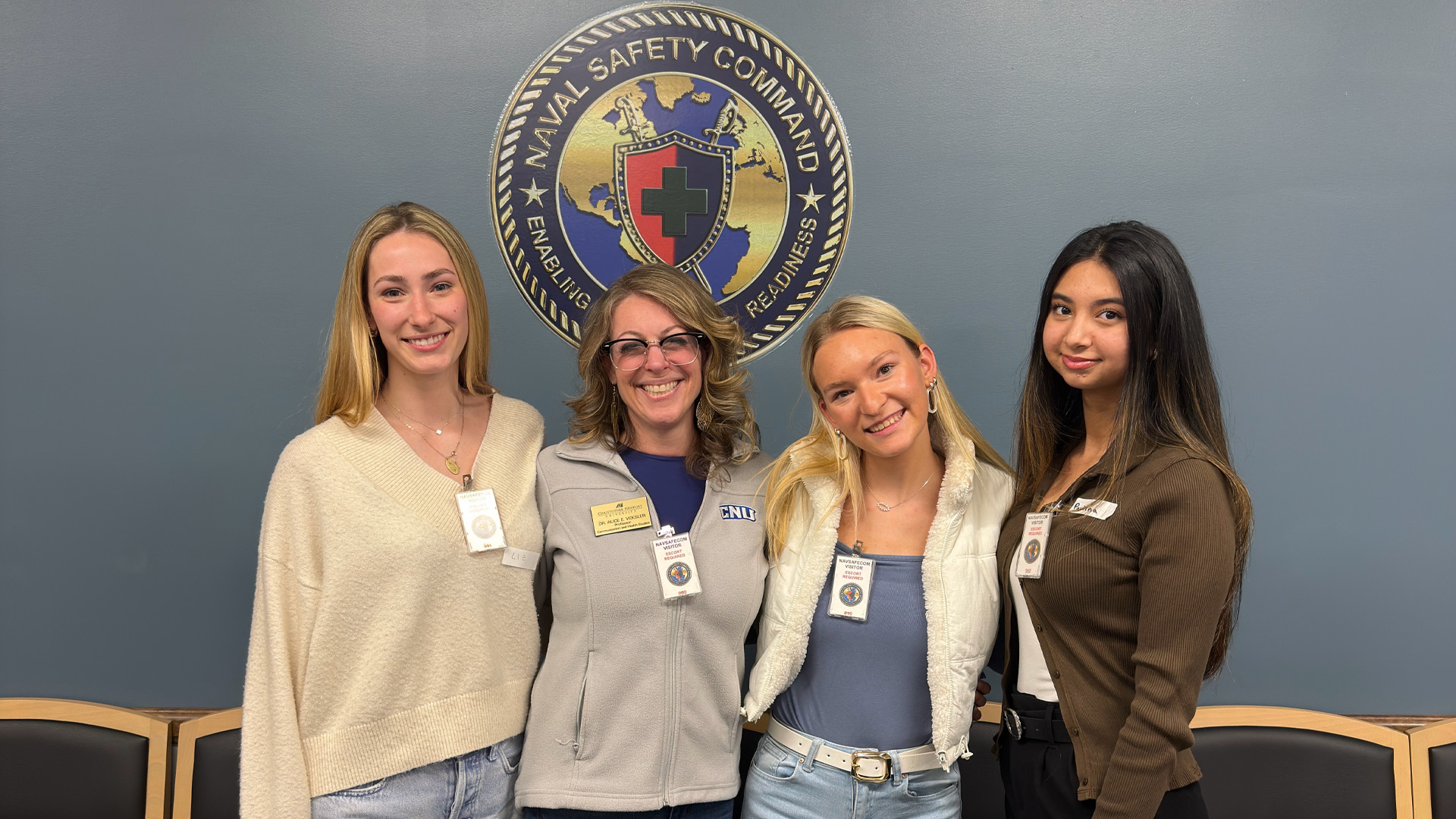 Students stand with their professor in front of the seal of Naval Safety Command seal on a wall.
