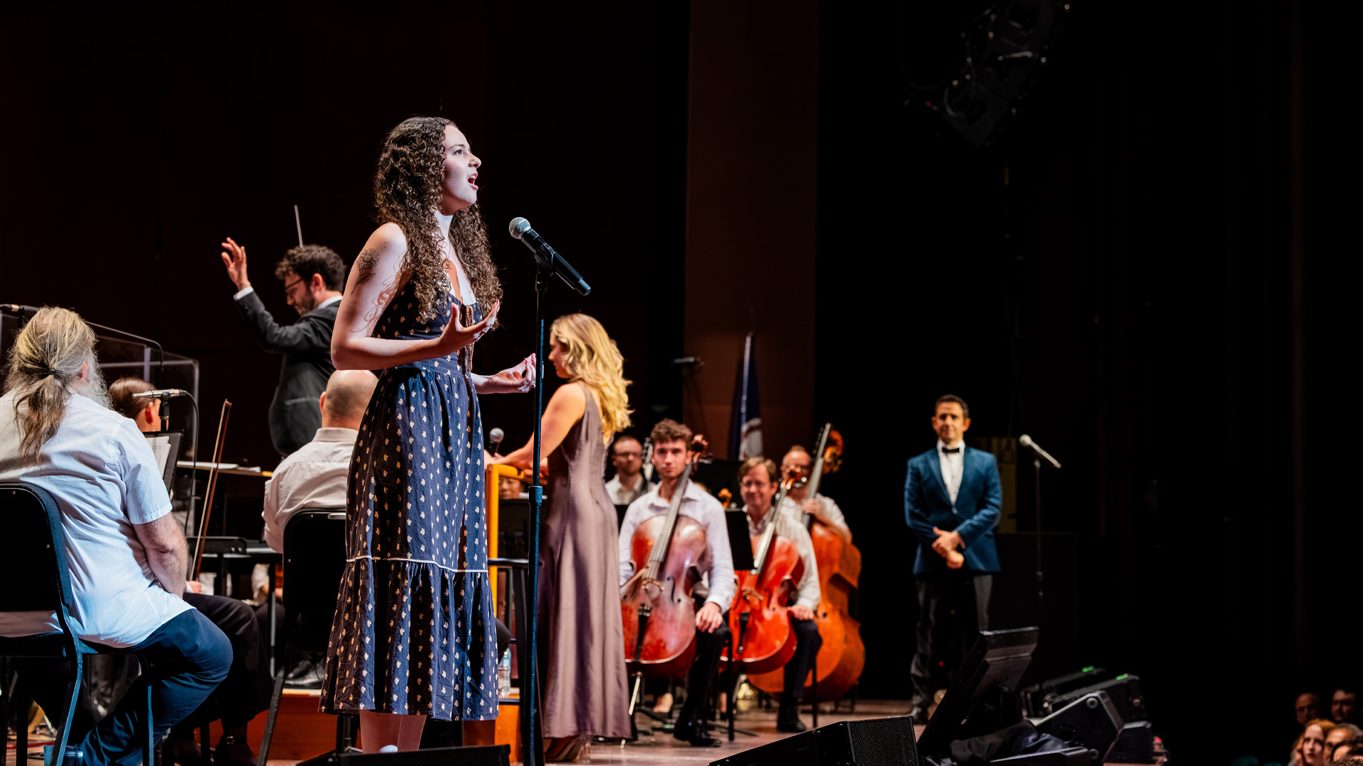 Erin Rigby in a polka dotted dress stands at a microphone and sings while an orchestra plays in the background