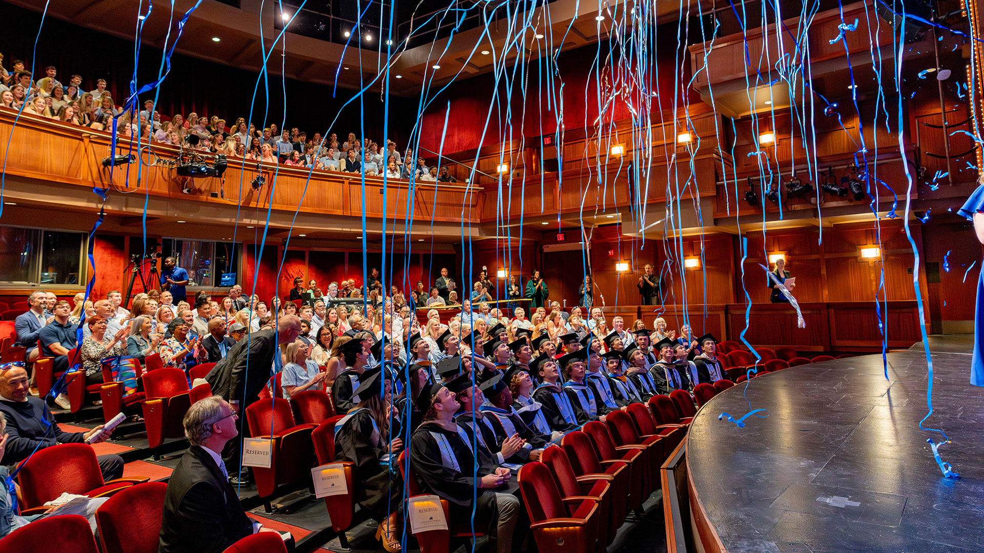 Student athletes and families look up as blue streamers fall from the ceiling during their commencement