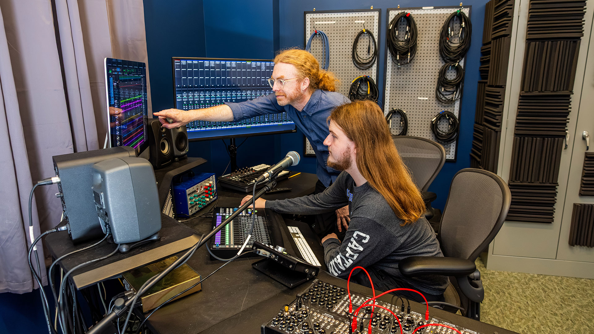 A professor points at a computer screen while a student is seated and looks on. They are in a music production studio