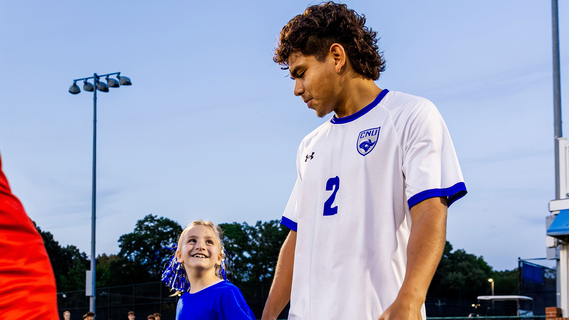 A male Christopher Newport University soccer player in a white and blue jersey looking down and smiling at a young girl in a blue shirt