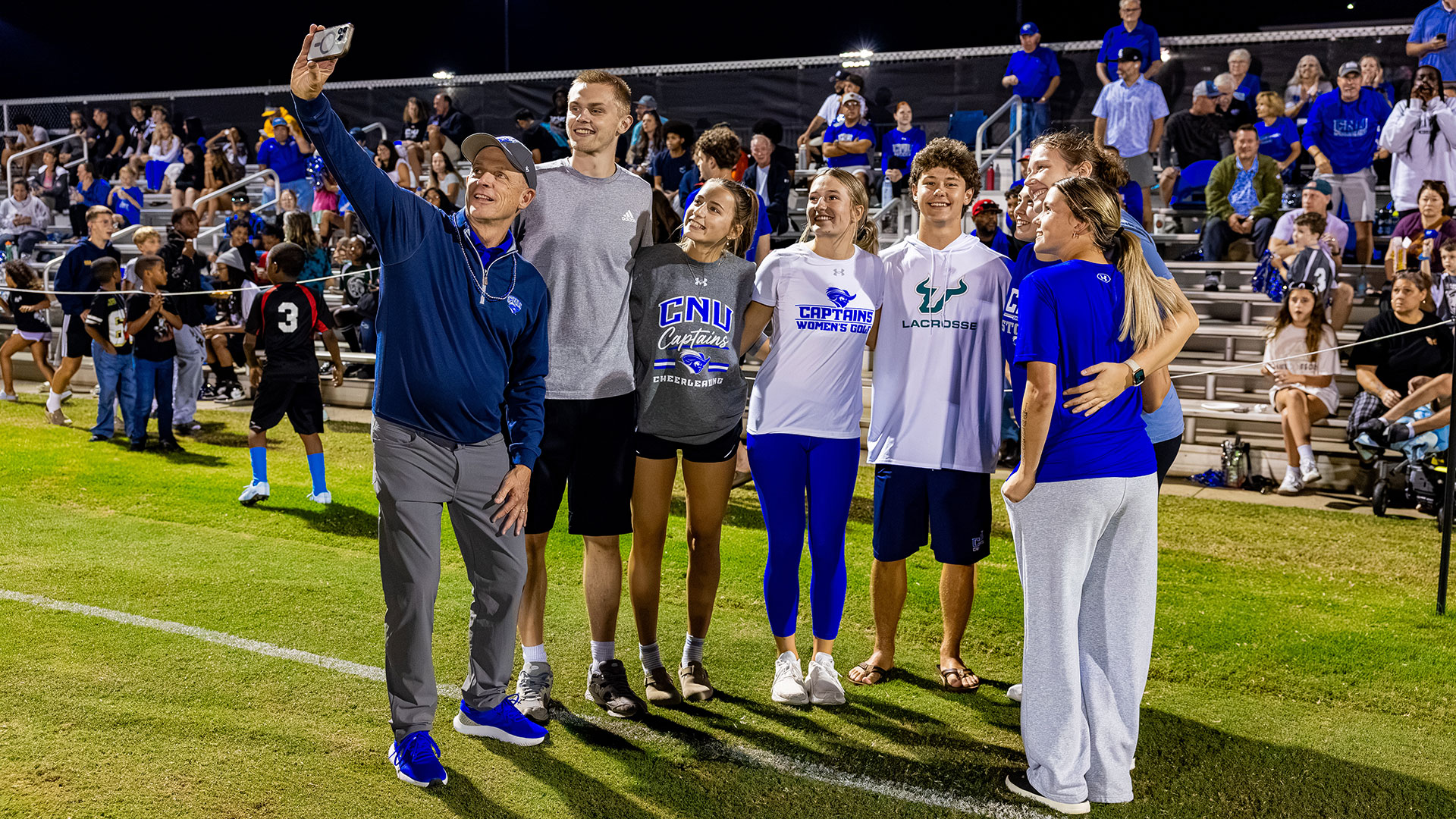 CNU President Bill Kelly holds up a smartphone to take a group selfie on an athletic field with several Christopher Newport University student-athletes